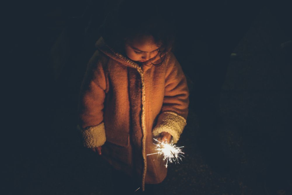 Little girl wearing brown jacket holding sparkler in her hand in the dark.