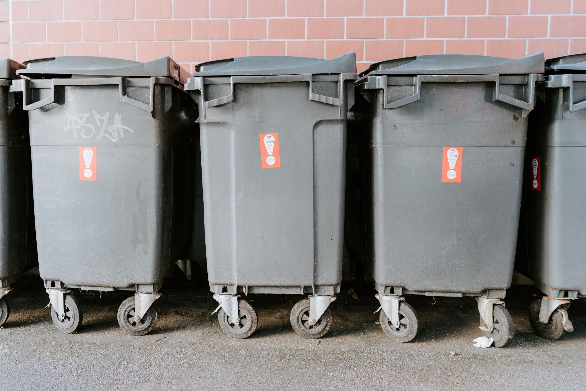 Dark grey industrial rubbish bins lined up side by side with red brick wall behind them.