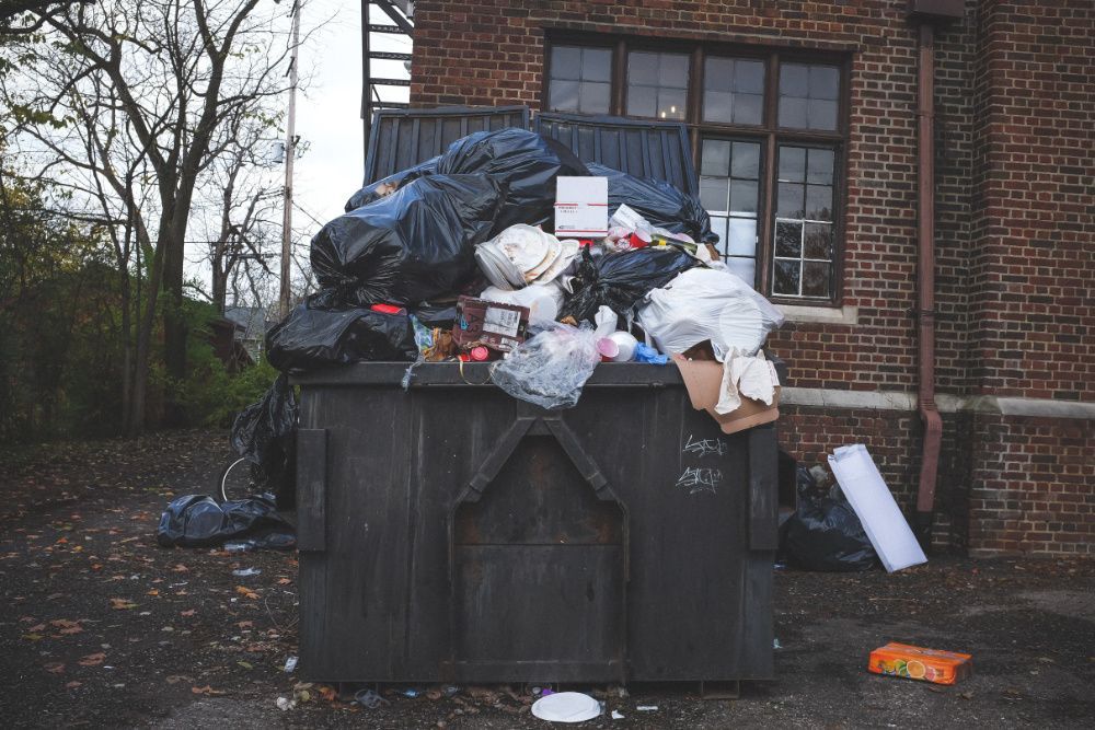 Black rubbish skip overflowing with rubbish bags in front of red brick building.