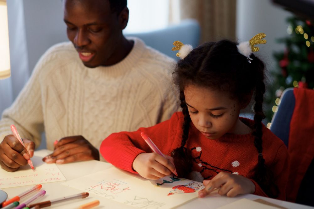 Man in white wooly sweater and daughter in red Christmas sweater drawing Santa Claus on Paper.