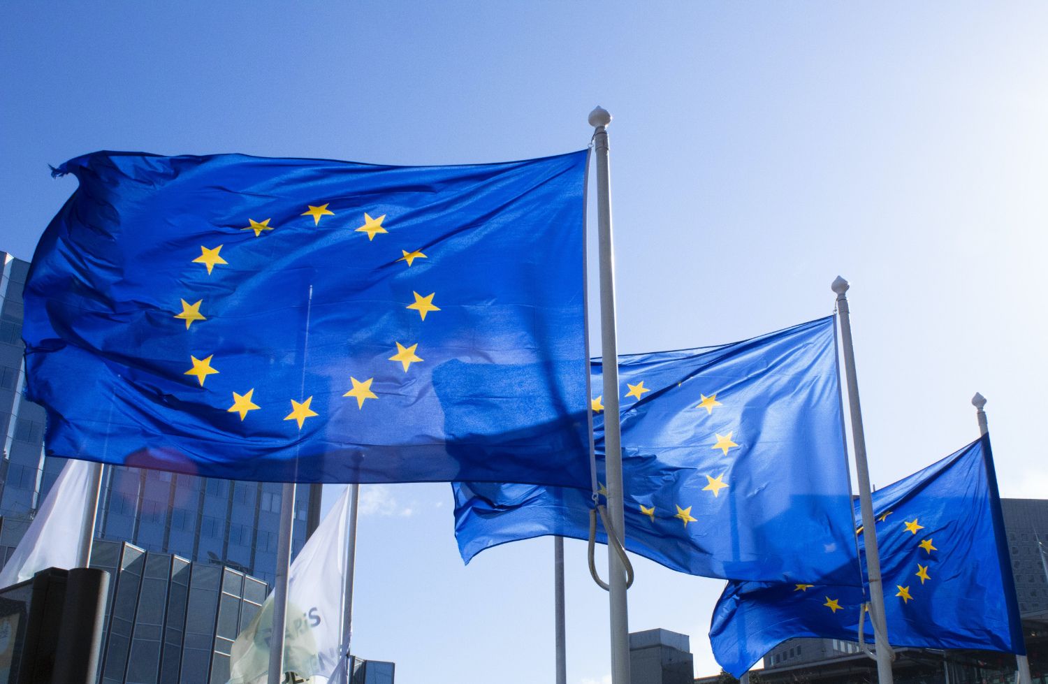 Three European union flags flying from right to left with blue sky and buildings in the background.