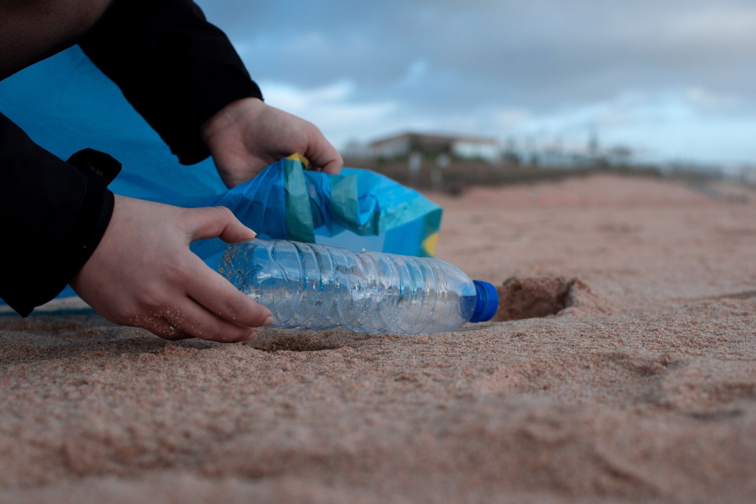 Person picking up plastic water bottle out of the send on a beach near the water.