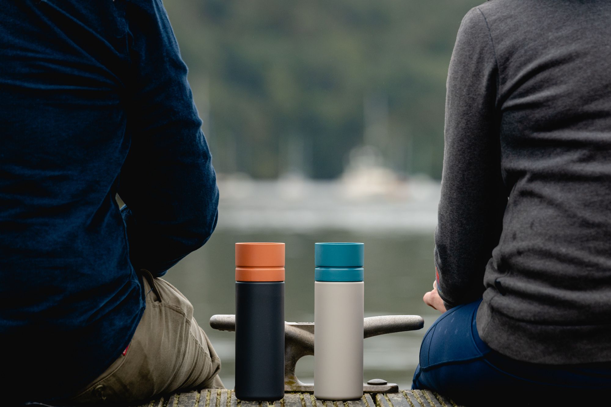 Two Circular&Co. reusable water bottles placed on wooden decking between two people sitting down facing a lake.