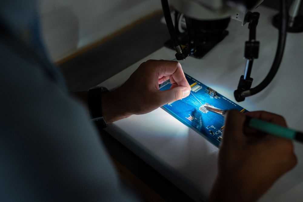 Person using tools to fix illuminated blue circuit board on white surface.