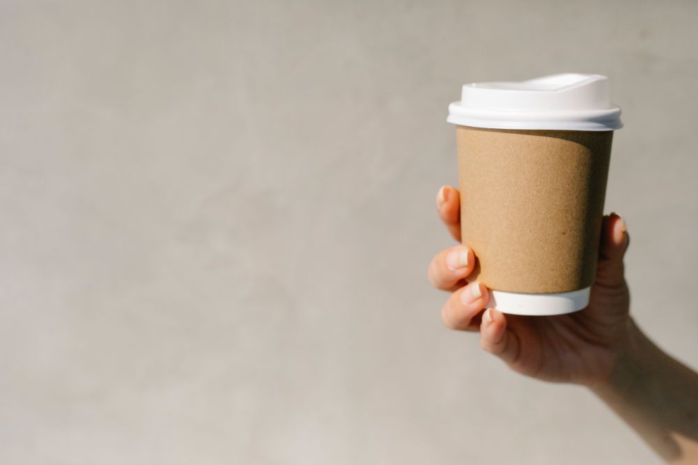 Brown and white single-life disposable coffee cup being held up against grey wall.