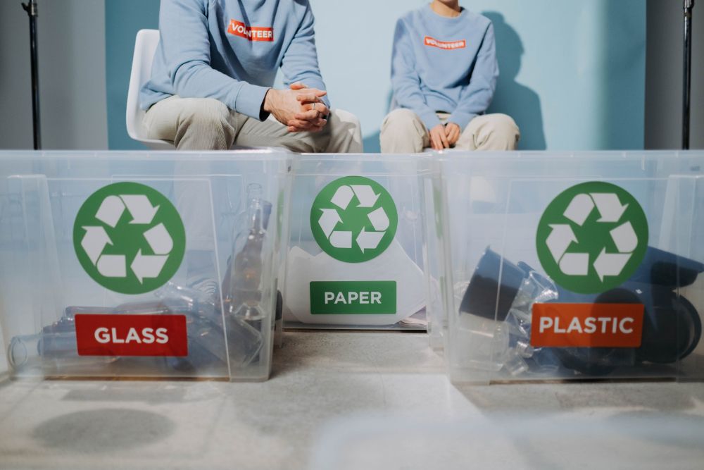 Two volunteers sorting glass, paper, and plastic items into clear boxes for recycling.