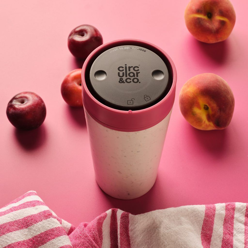 white reusable cup with pink lid in front of pink fruit on a pink table