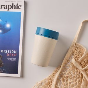 white reusable cup with blue lid on table with magazine and tote bag