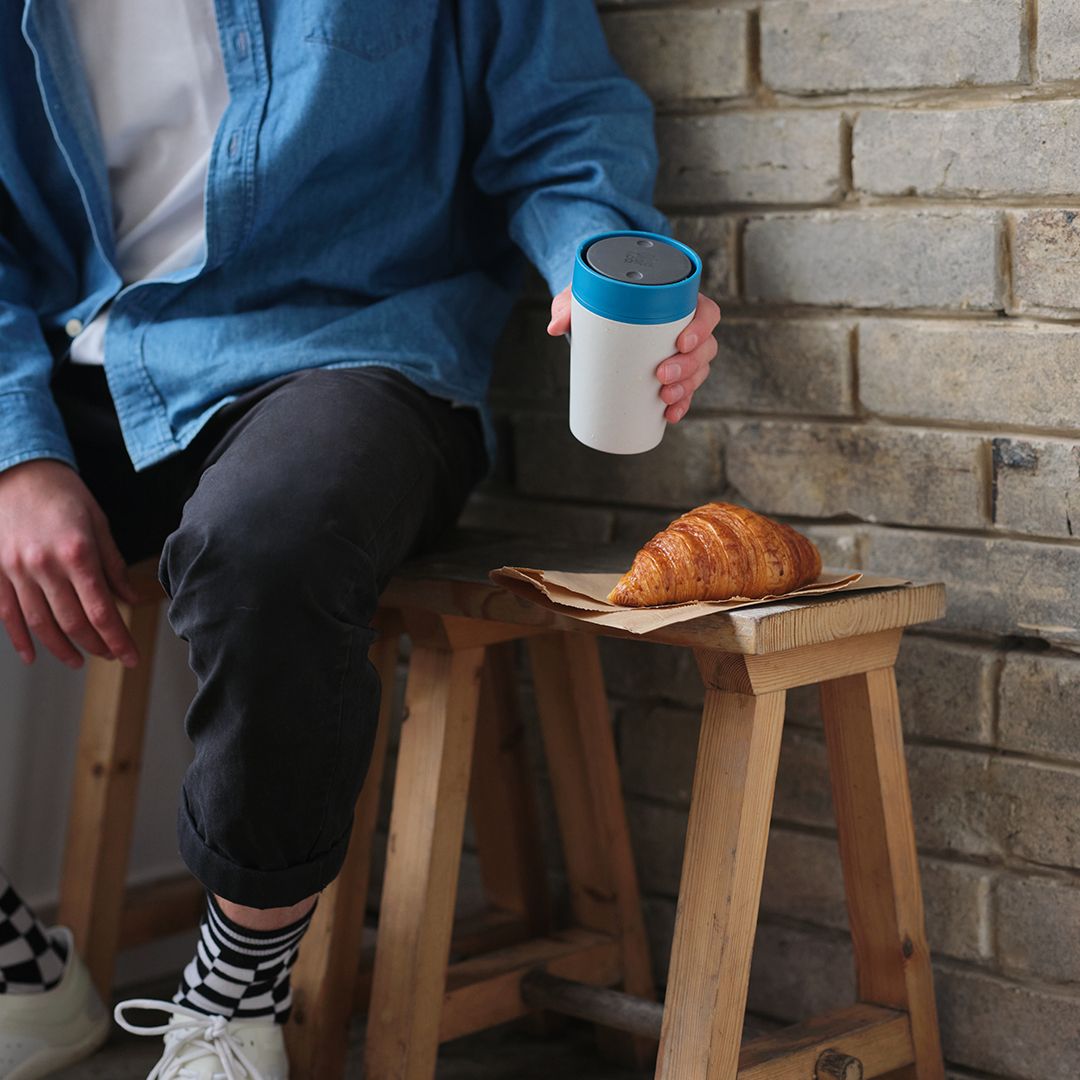 man holding white reusable cup with blue lid on a bench