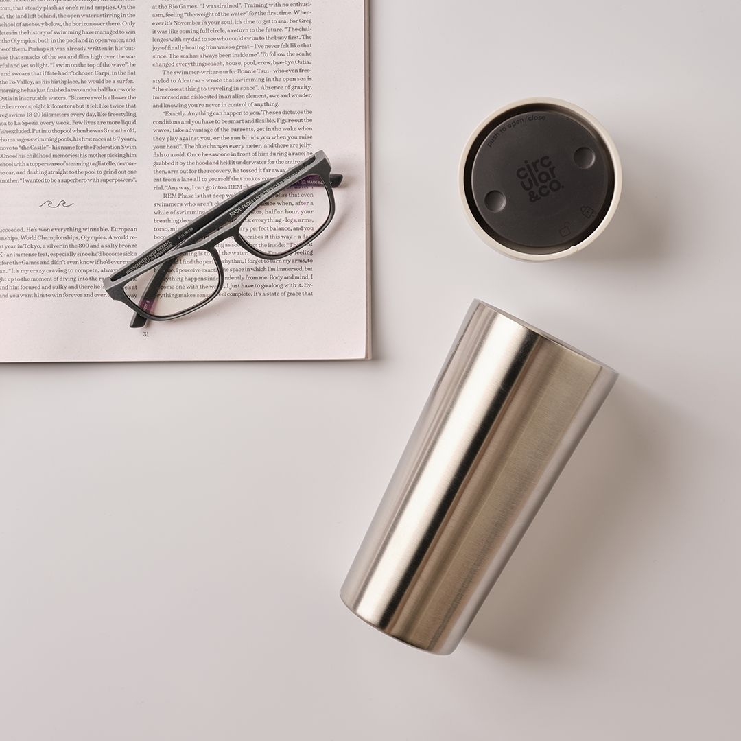 steel reusable cup on its side next to glasses and newspaper