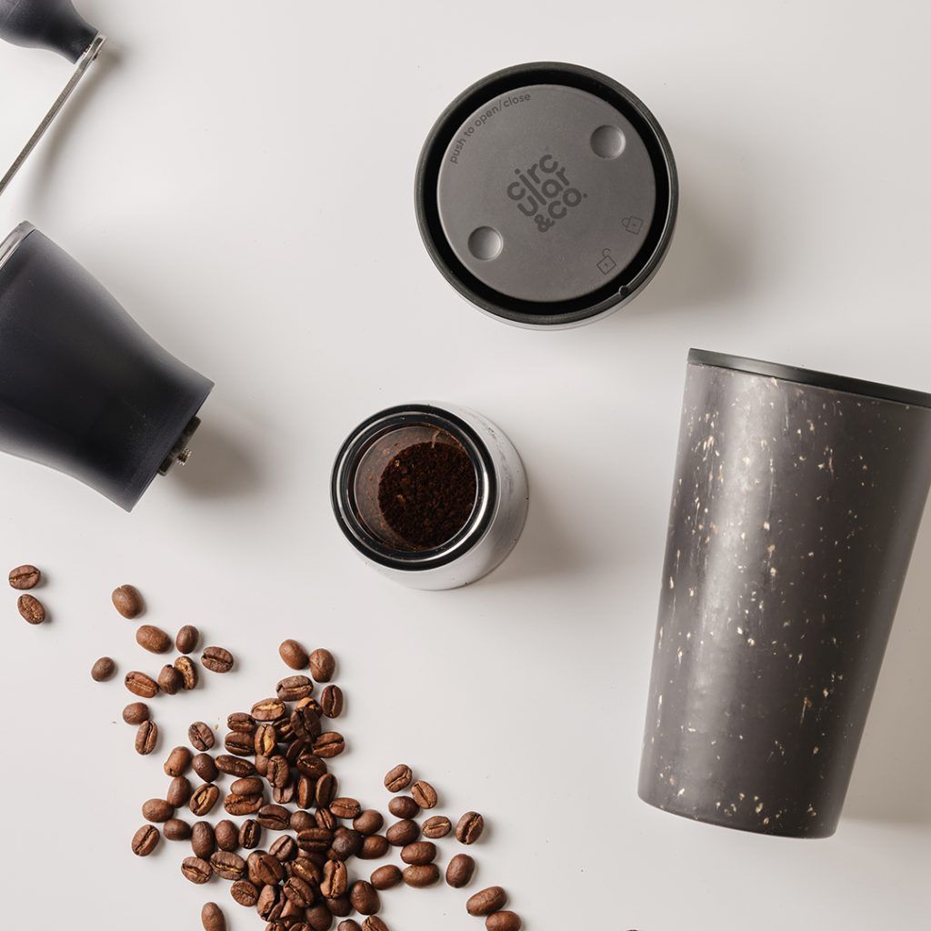 black reusable cup on white table with coffee beans and black lid