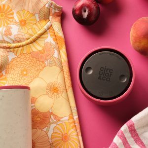 pink reusable cup lid on a pink table next to a colourful wetsuit and pink fruit