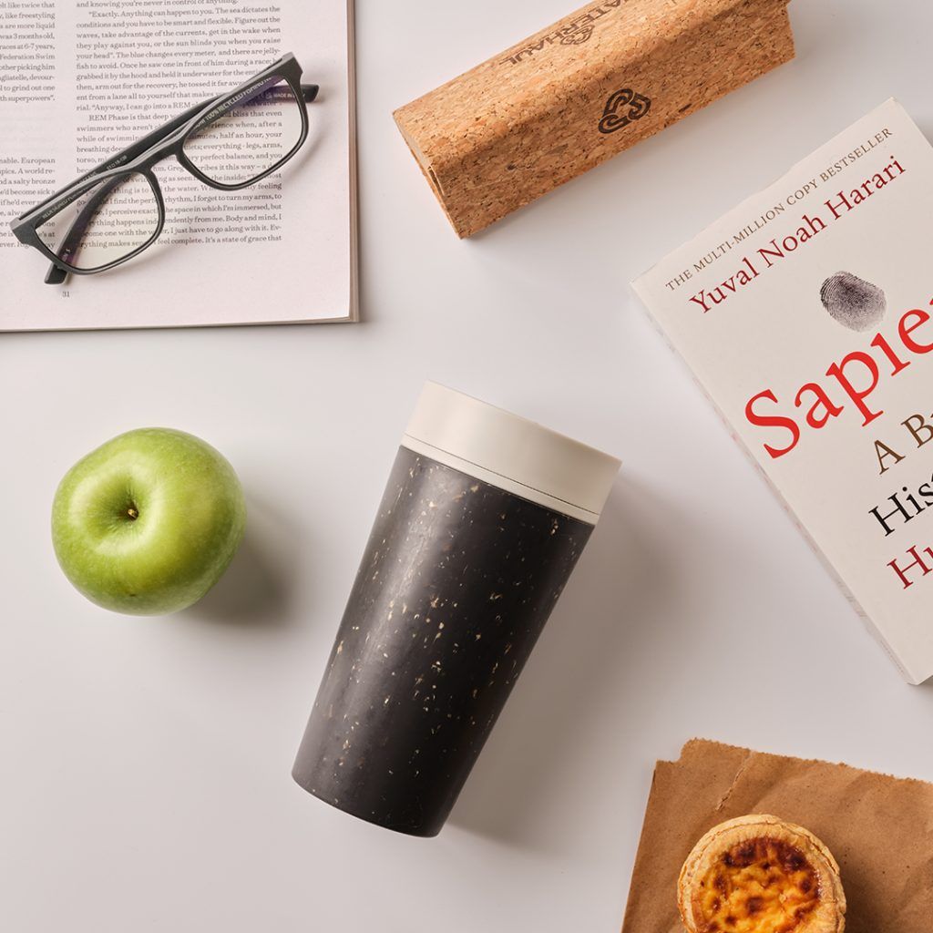 black reusable cup with white lid on table along with apple, books and sunglasses case