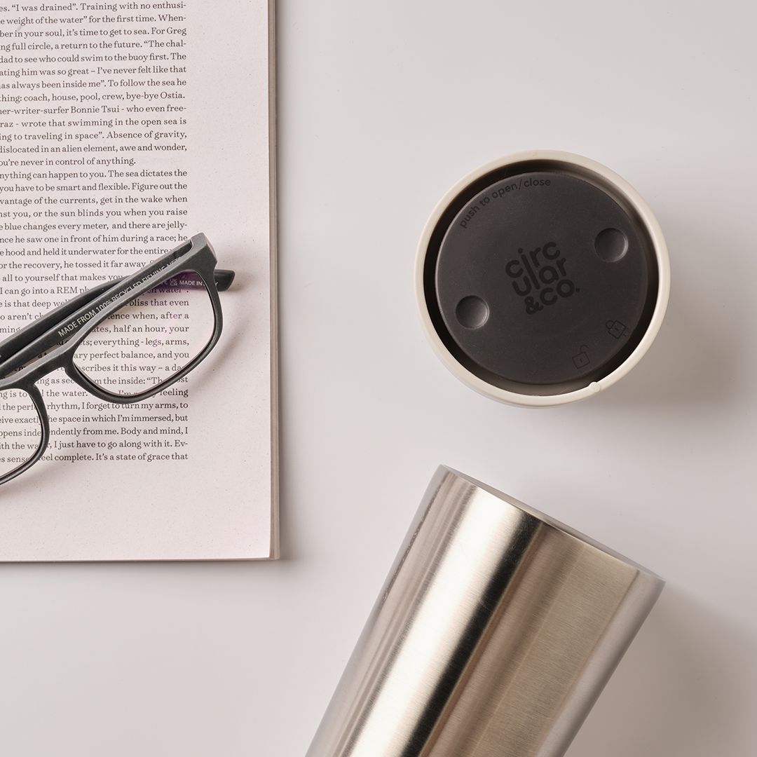 steel reusable cup with white lid next to glasses and book