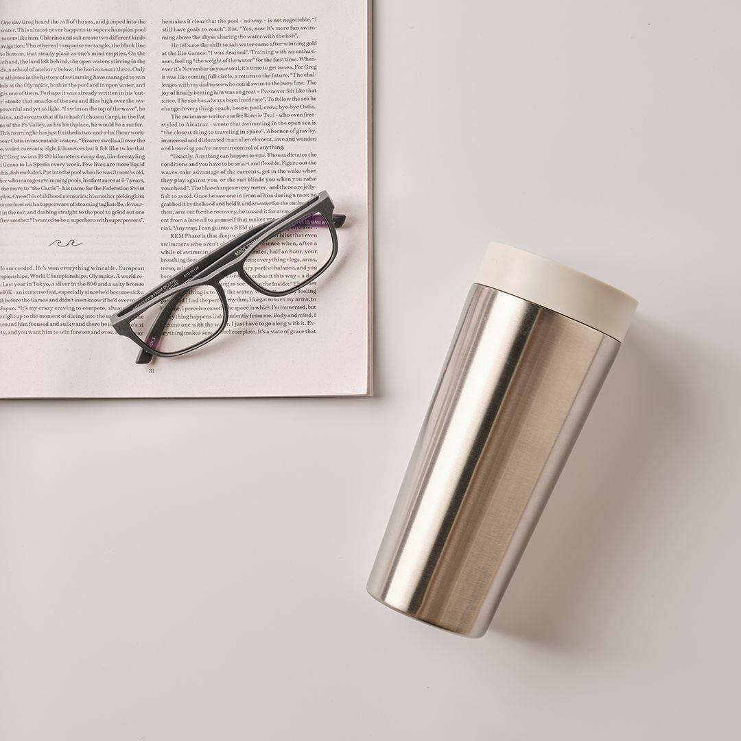 steel reusable cup with white lid next to newspaper and glasses