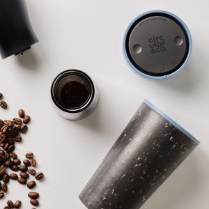 black reusable cup with blue lid on white table with coffee beans