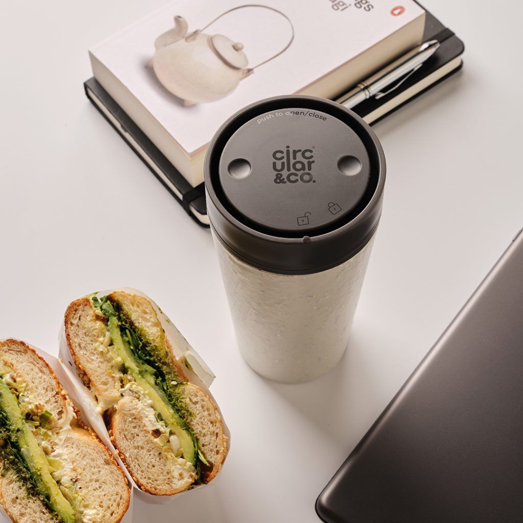 white reusable cup with black lid on table with bagel and books