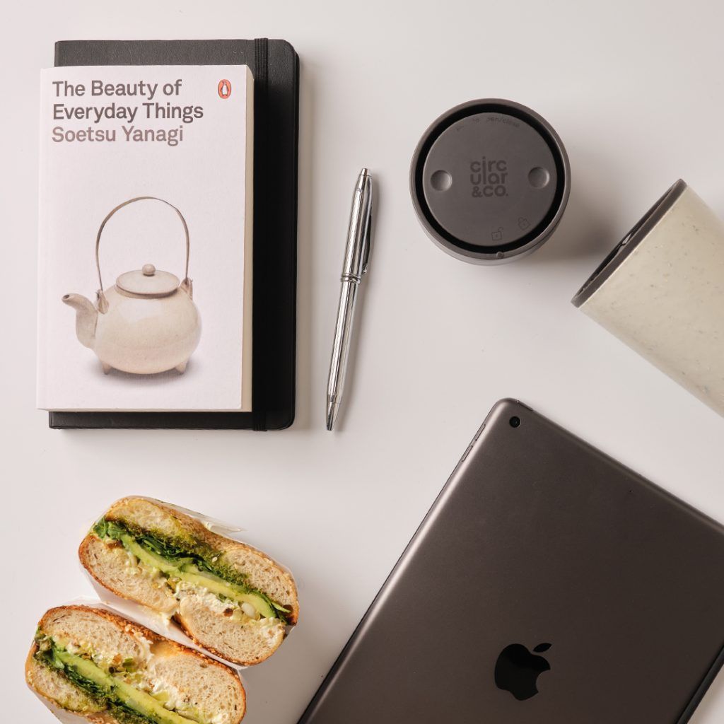 collection of reusable cup, ipad, bagel and book on a table