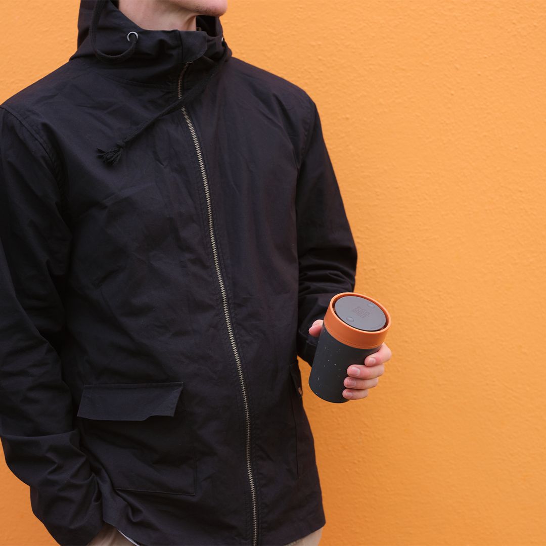 man holding black reusable cup with orange lid in front of an orange wall
